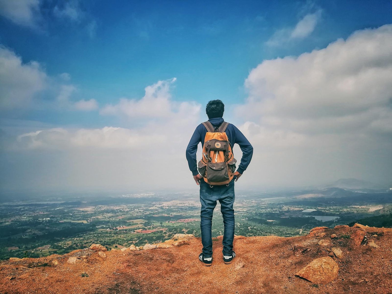 man in blue dress shirt and blue jeans and orange backpack standing on mountain cliff looking at town under blue sky and white clouds
